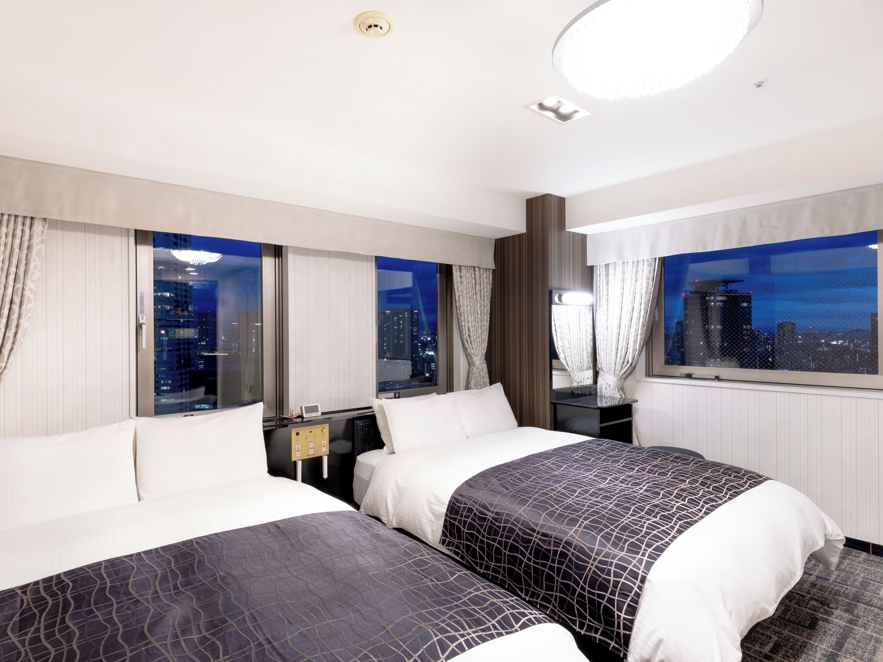 A hotel room with two beds, a nightstand, a mirror, and a view of the cityscape at night.