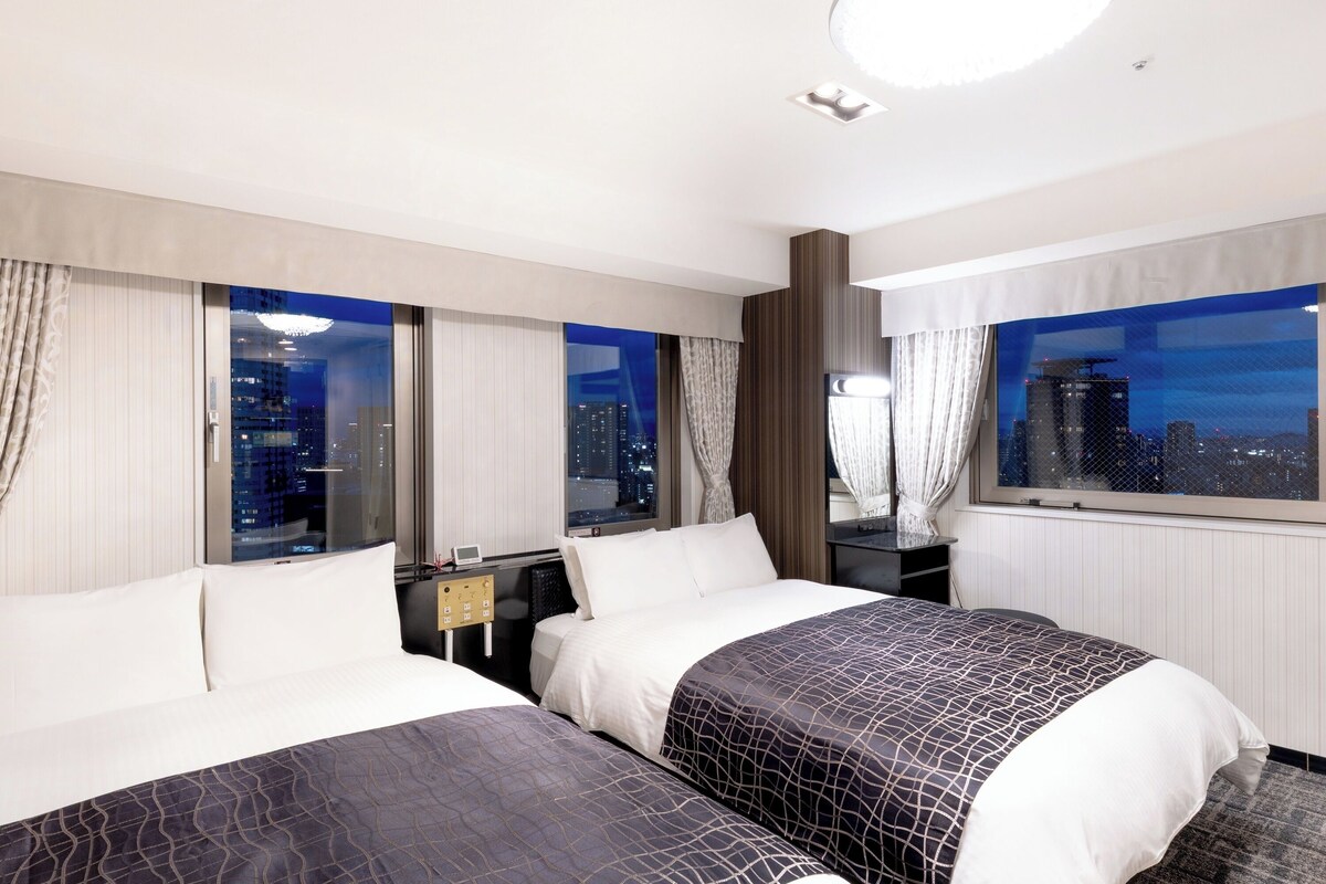 Habitación de hotel con dos camas, una mesita de noche, un espejo y vistas a la ciudad de noche.