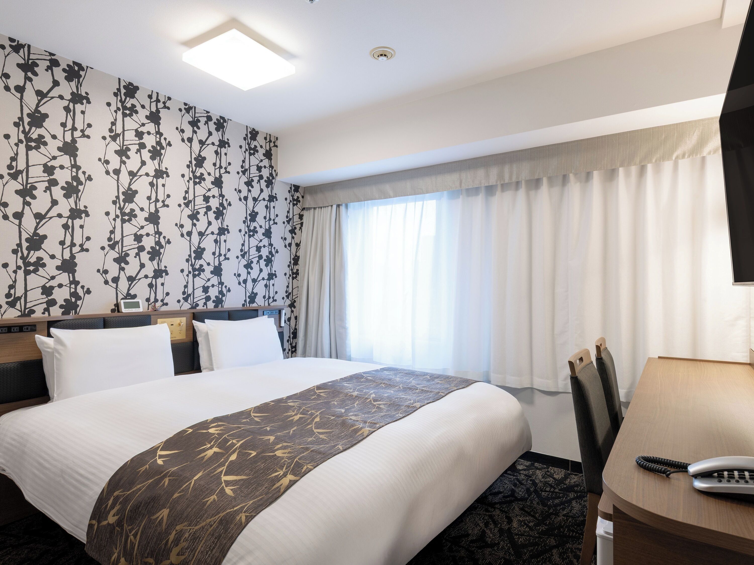 Ett hotellrum med en säng, ett skrivbord med en telefon, ett fönster med gardiner och en vägg med ett blommönster.