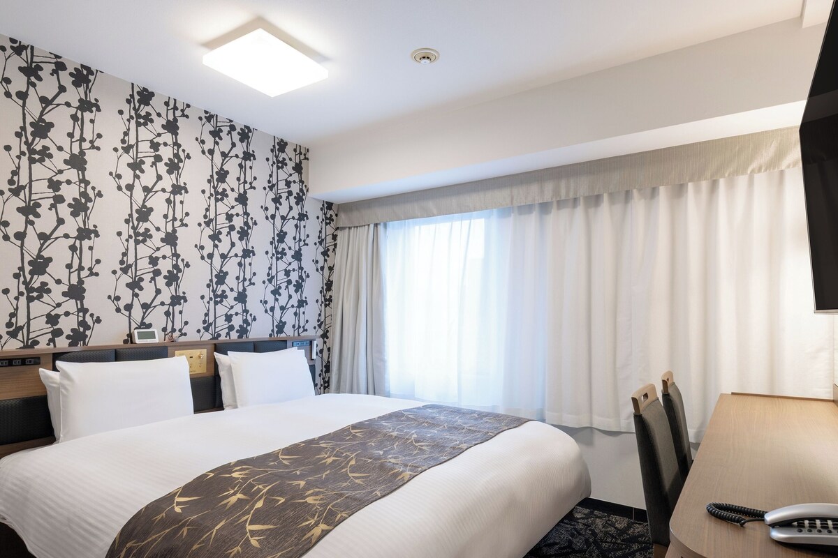 Habitación de hotel con una cama, un escritorio con teléfono, una ventana con cortinas y una pared con estampado floral.