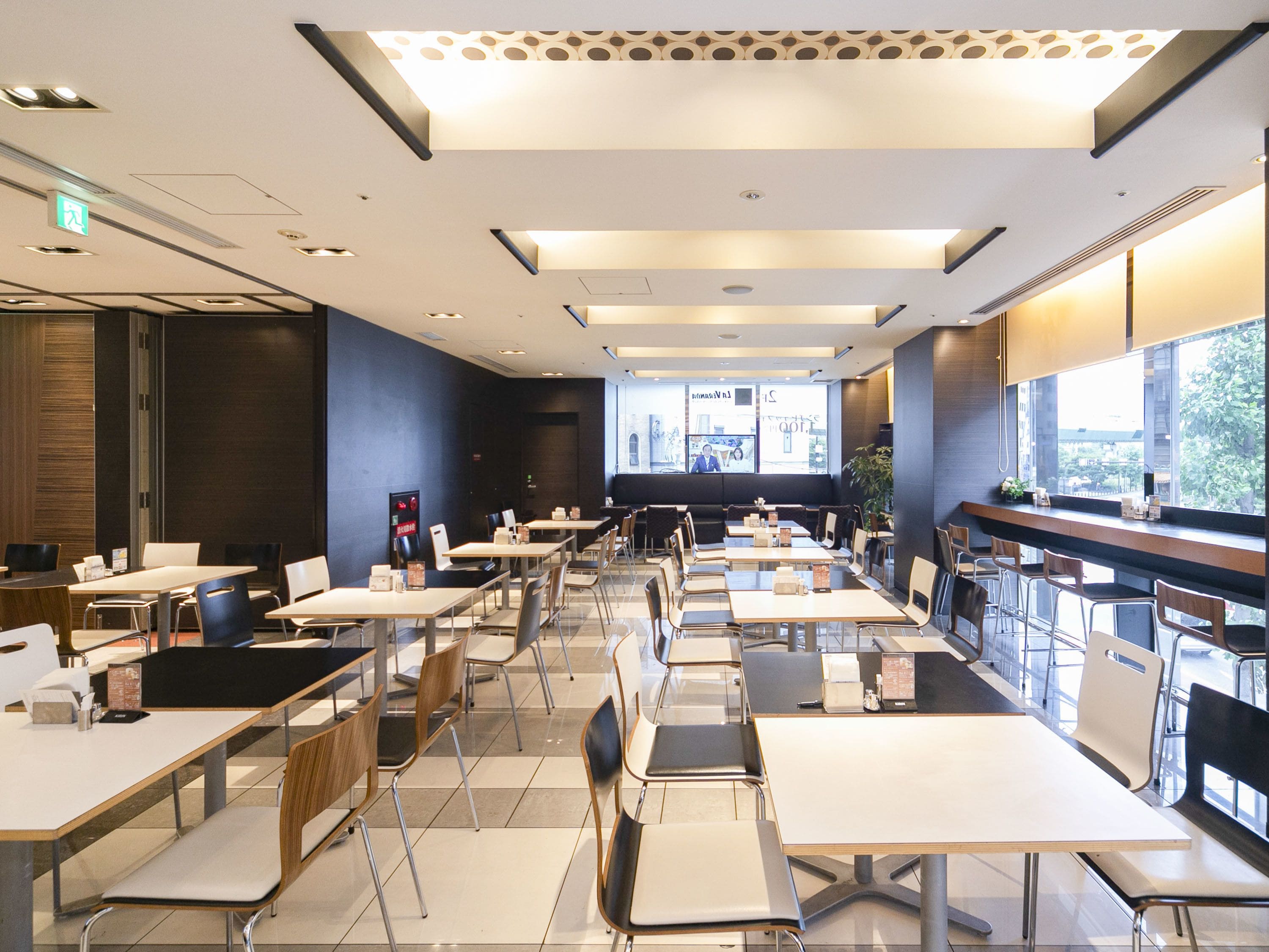 A modern restaurant interior with multiple dining tables and chairs, a bar area, and large windows providing natural light.
