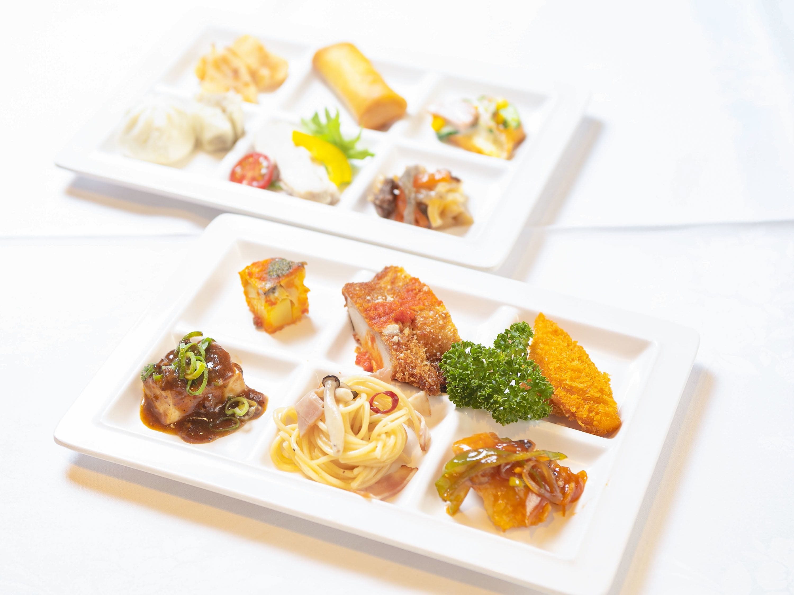Two square white plates with various dishes including pasta, a piece of meat, and vegetables.
