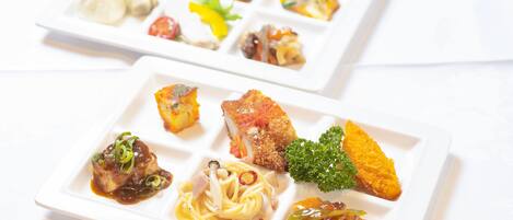 Two square white plates with various dishes including pasta, a piece of meat, and vegetables.