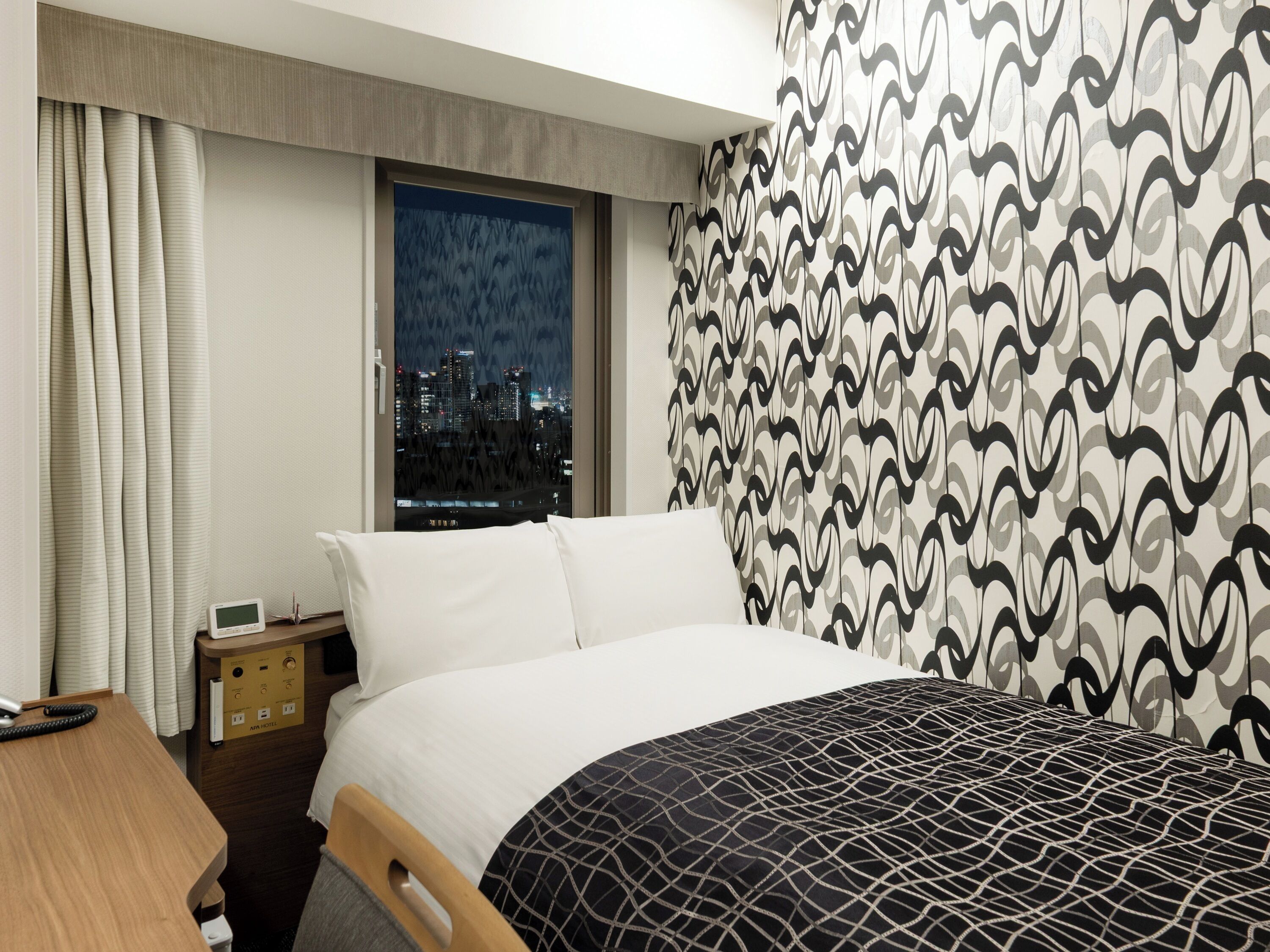 A hotel room with a bed, a desk, a nightstand, and a view of a cityscape at night.