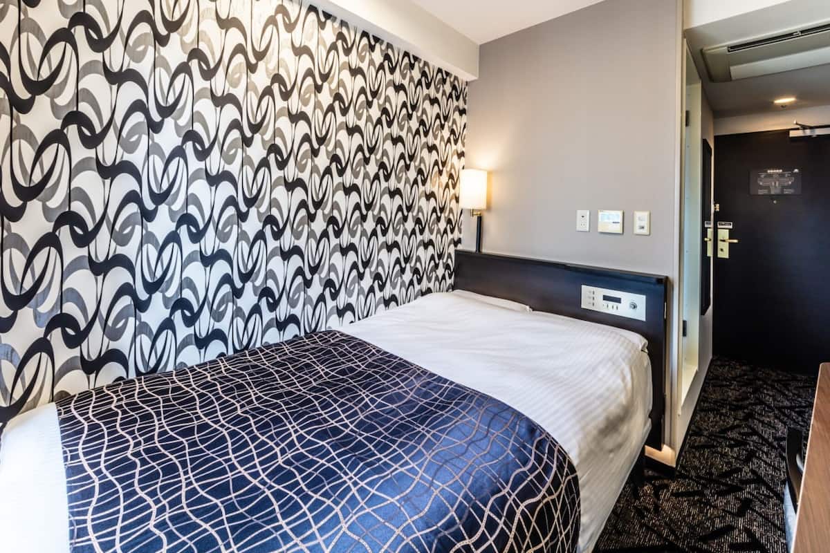 Habitación de hotel con pared de diseño, cama con colcha de motivos geométricos, mesita de noche y lámpara.