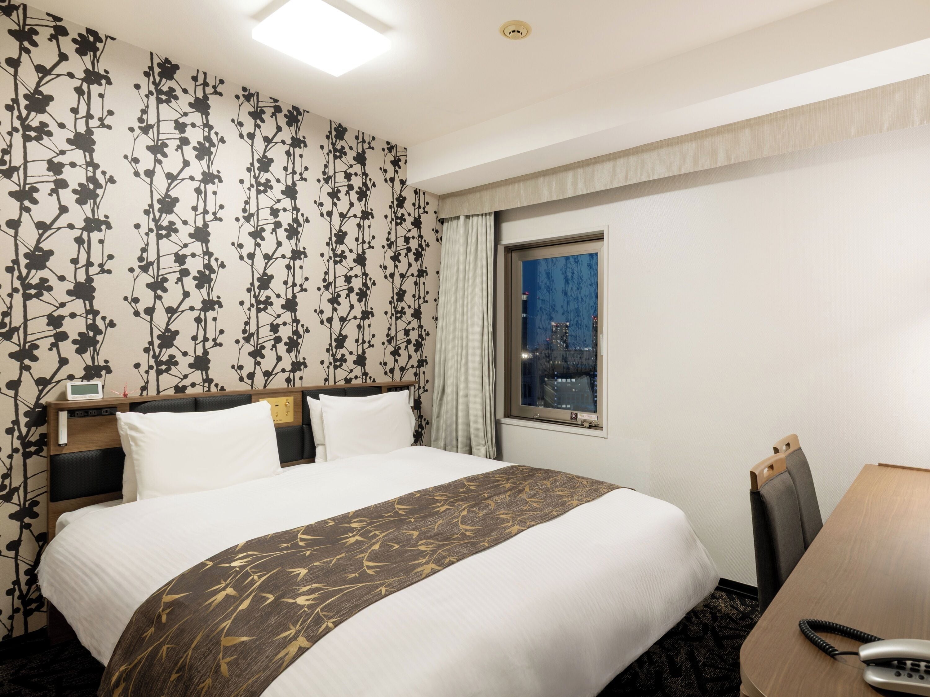 A hotel room with a bed, a desk with a telephone, a window with a city view, and a wall with a floral pattern.