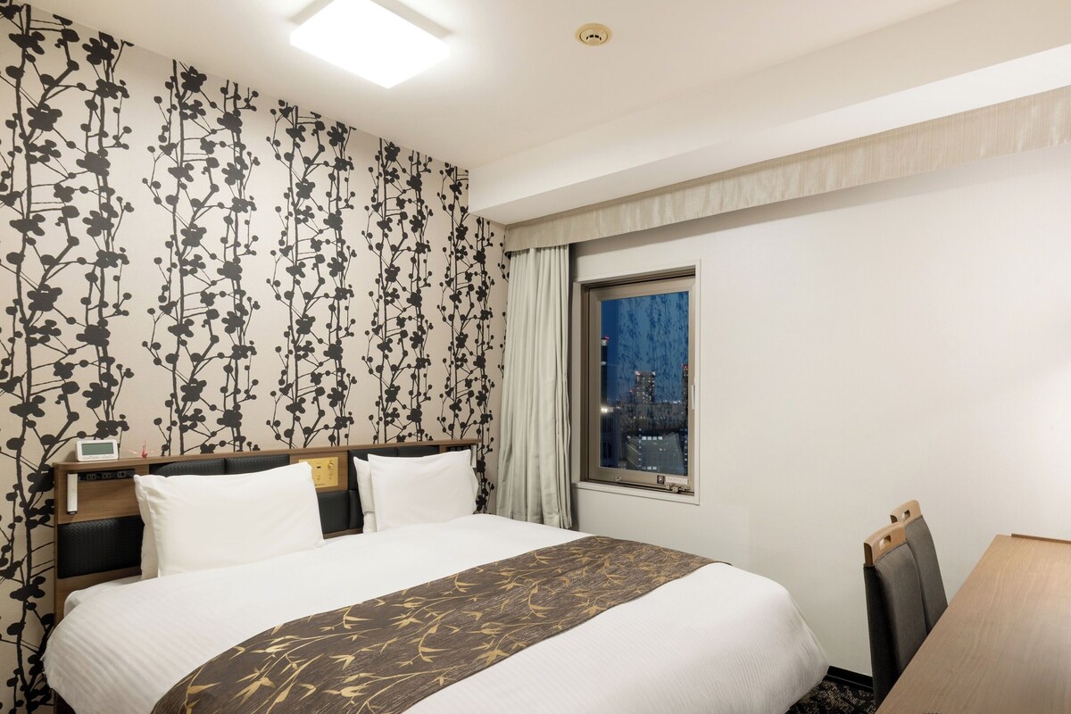 Habitación de hotel con una cama, un escritorio con teléfono, una ventana con vista a la ciudad y una pared con estampado floral.