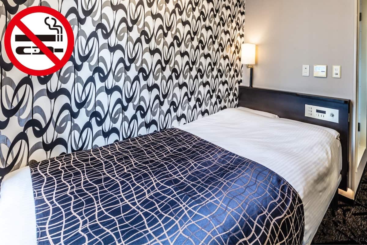 Habitación de hotel con pared estampada, cama con colcha azul y blanca, televisor montado en la pared y cartel de prohibido fumar.