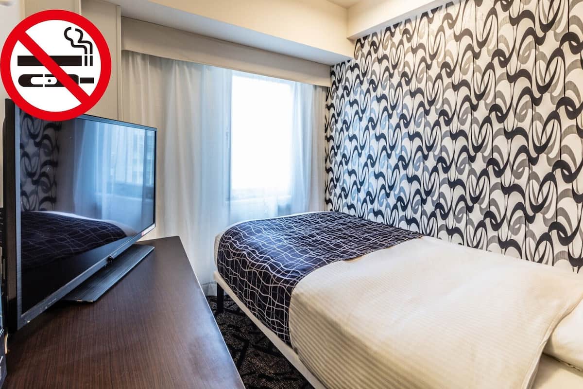 Una habitación de hotel con cama, televisor sobre un mueble, cartel de prohibido fumar y papel tapiz estampado.