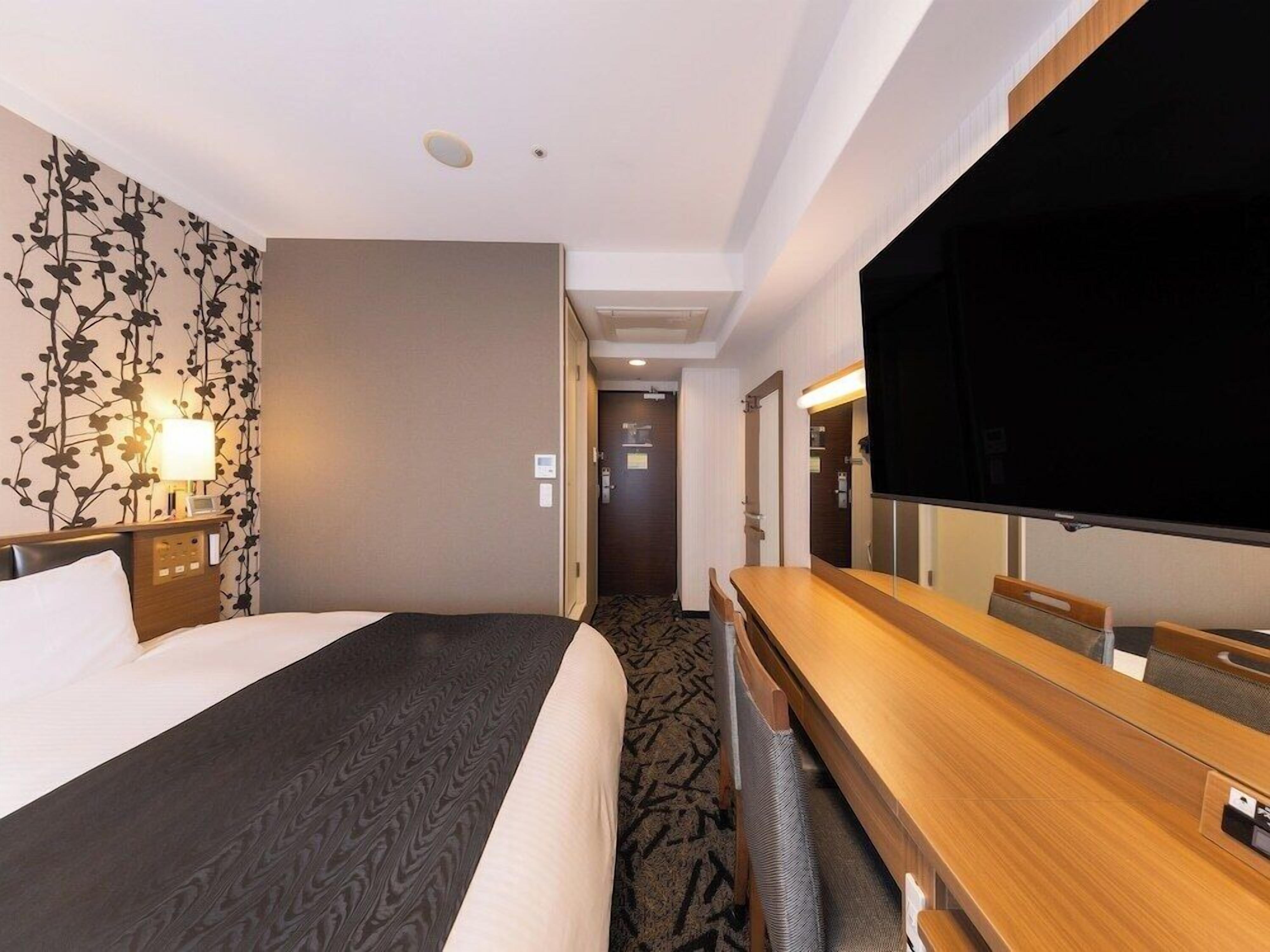 A hotel room with a bed, a desk with a flat-screen TV, a mirror, a wall-mounted lamp, and a carpeted floor.