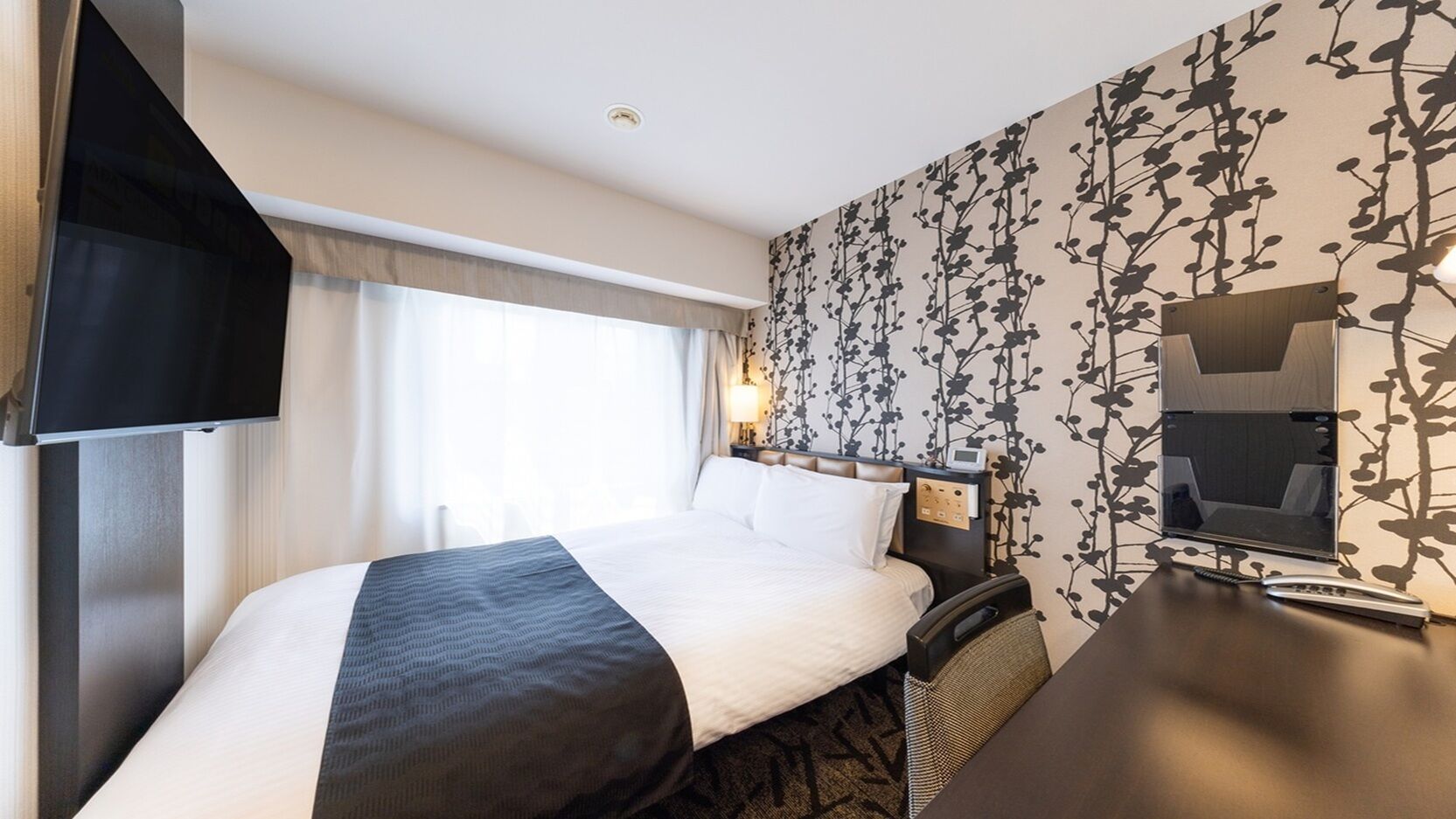 Habitación de hotel con cama, escritorio, televisor y una pared con estampado floral.