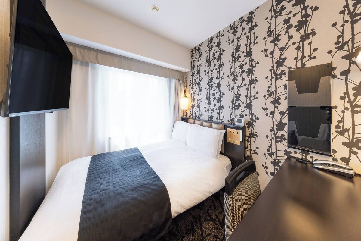 A hotel room with a bed, a desk with a telephone, a television, and a window with curtains.