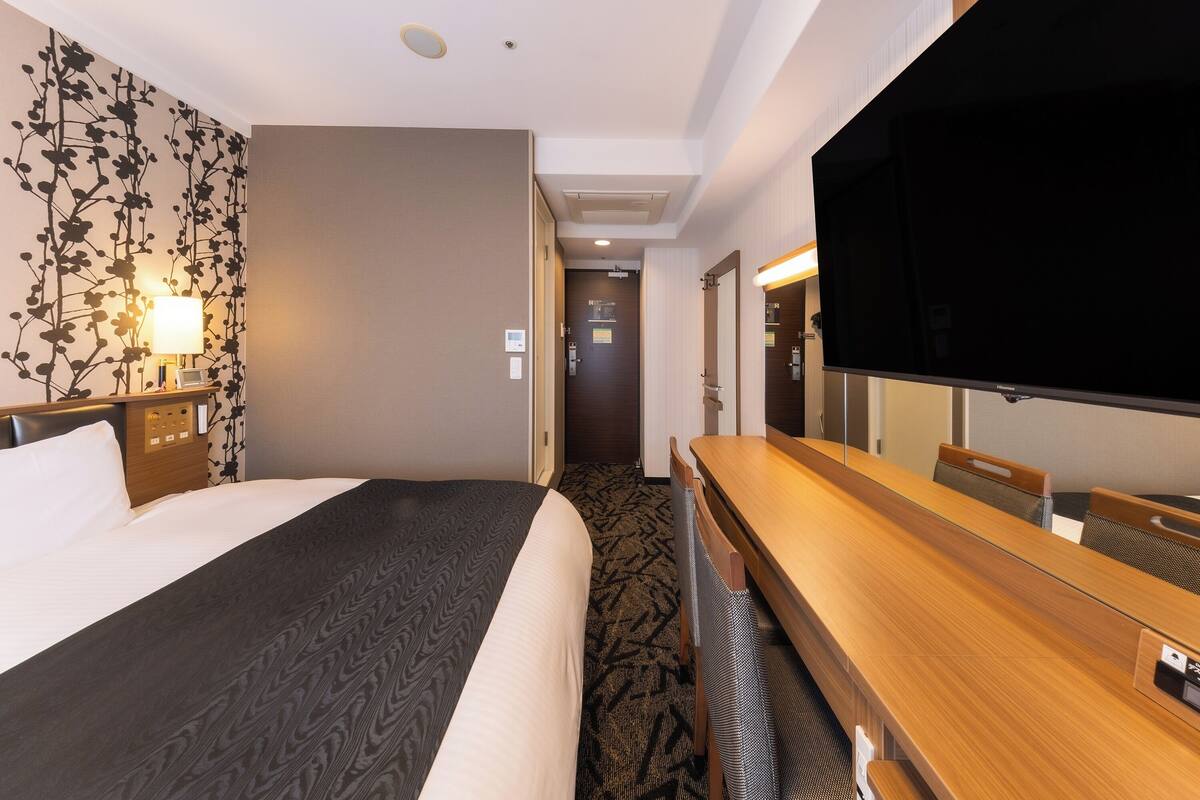 A hotel room with a bed, a desk, a flat-screen TV, and a wall-mounted lamp.