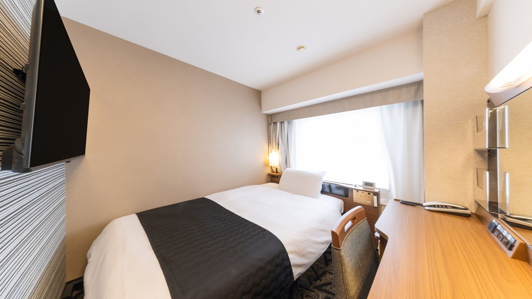 Habitación de hotel con cama, escritorio, silla, televisor, ventana con cortinas y panel de control en la pared.