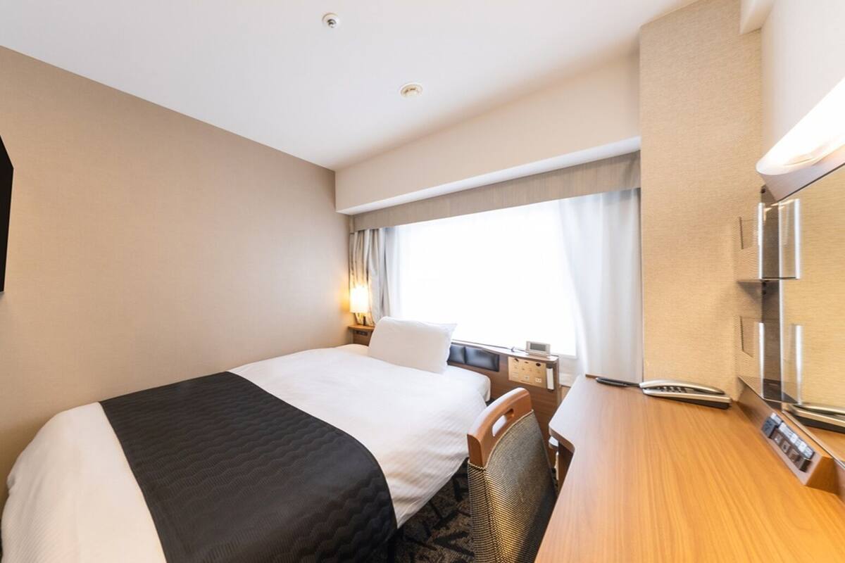 A hotel room with a bed, a desk, a chair, a TV, a window with curtains, and a wall-mounted control panel.