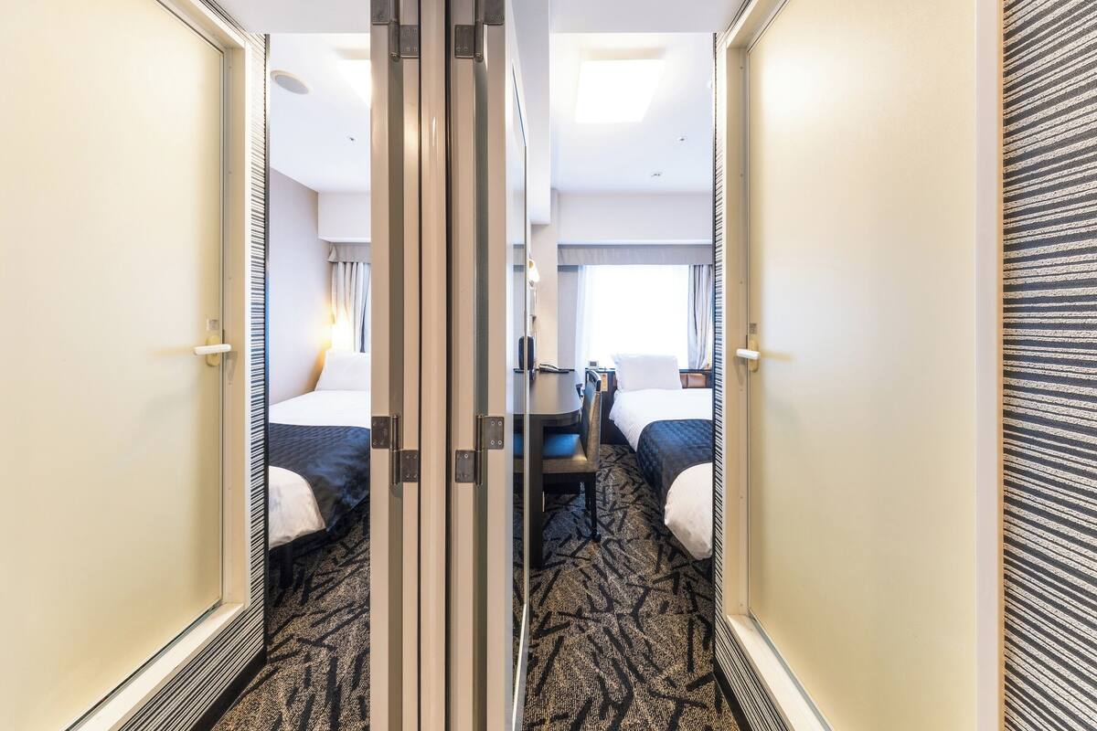 A hotel room with a mirrored closet door, a bed with a white comforter, a bedside table, and a window with curtains.