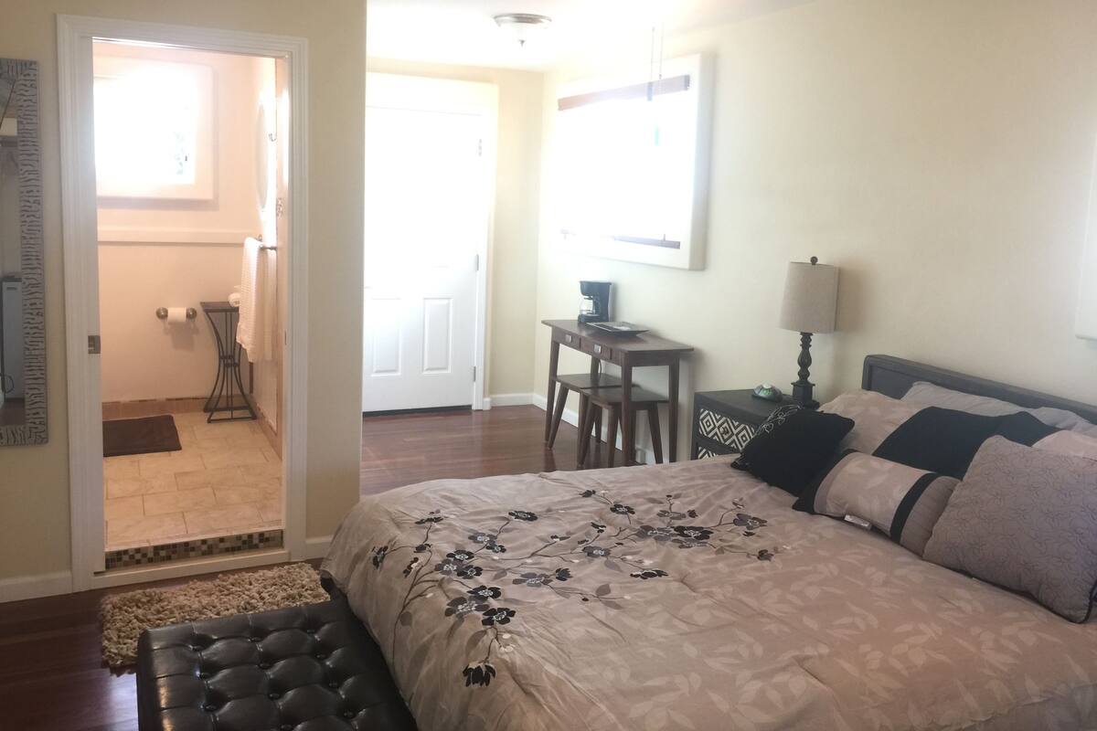 A bedroom with a large bed, a desk, a chair, a lamp, and a view of the outside.