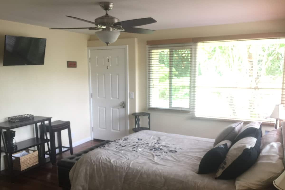 A bedroom with a bed, a ceiling fan, a TV mounted on the wall, a window with blinds, and a small table with a basket.