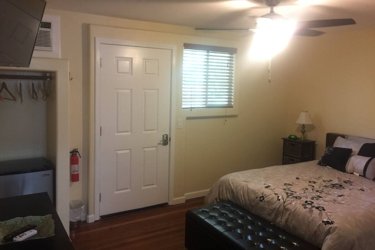 A bedroom with a bed, a ceiling fan, a window with blinds, a nightstand with a lamp, and a closet with clothes and a fire extinguisher.