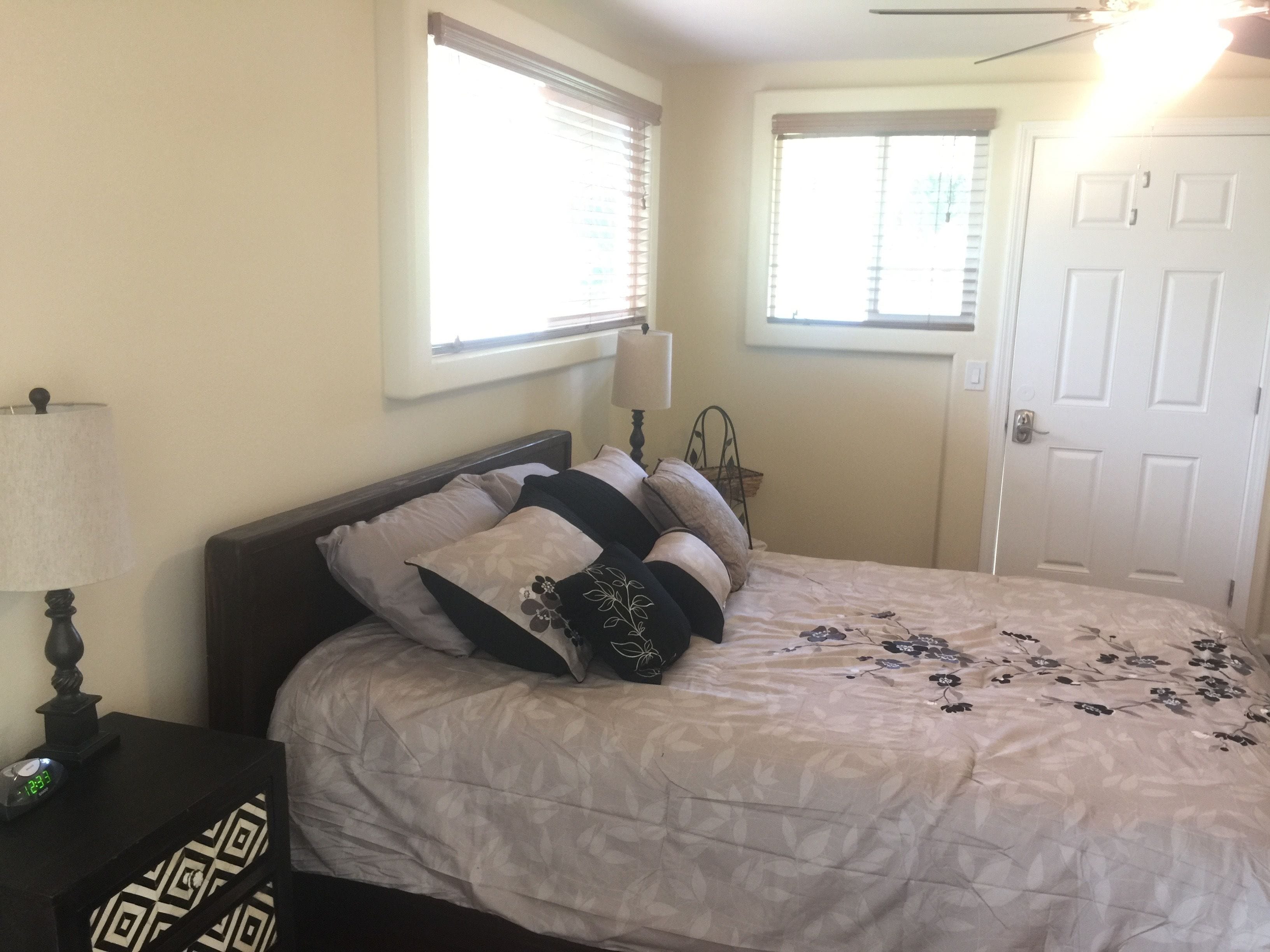 A bedroom with a bed, a nightstand with a lamp, and two windows with blinds.