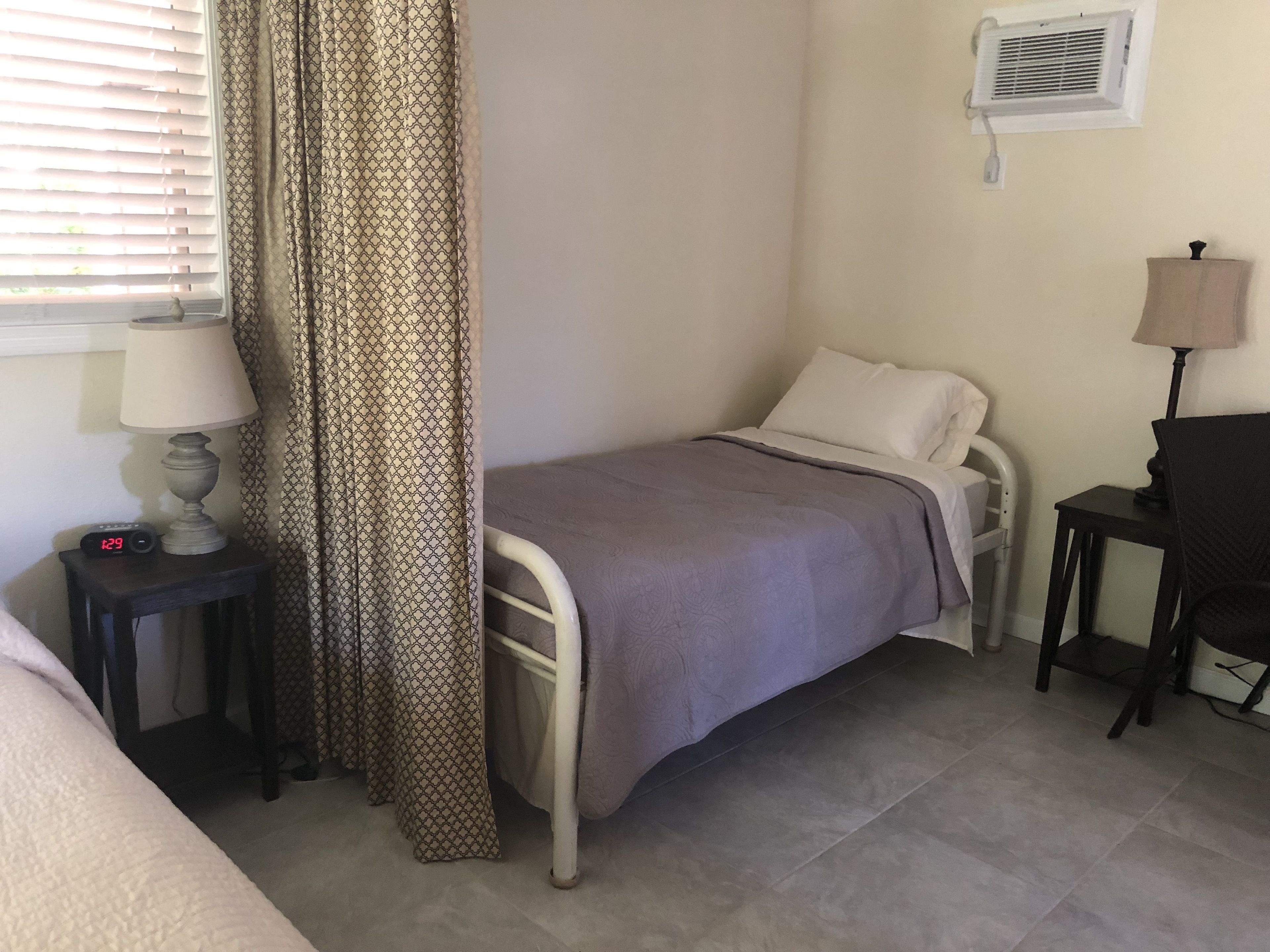 Un dormitorio pequeño y ordenado con una cama individual, una mesita de noche con lámpara, una silla y un aire acondicionado instalado en la pared.