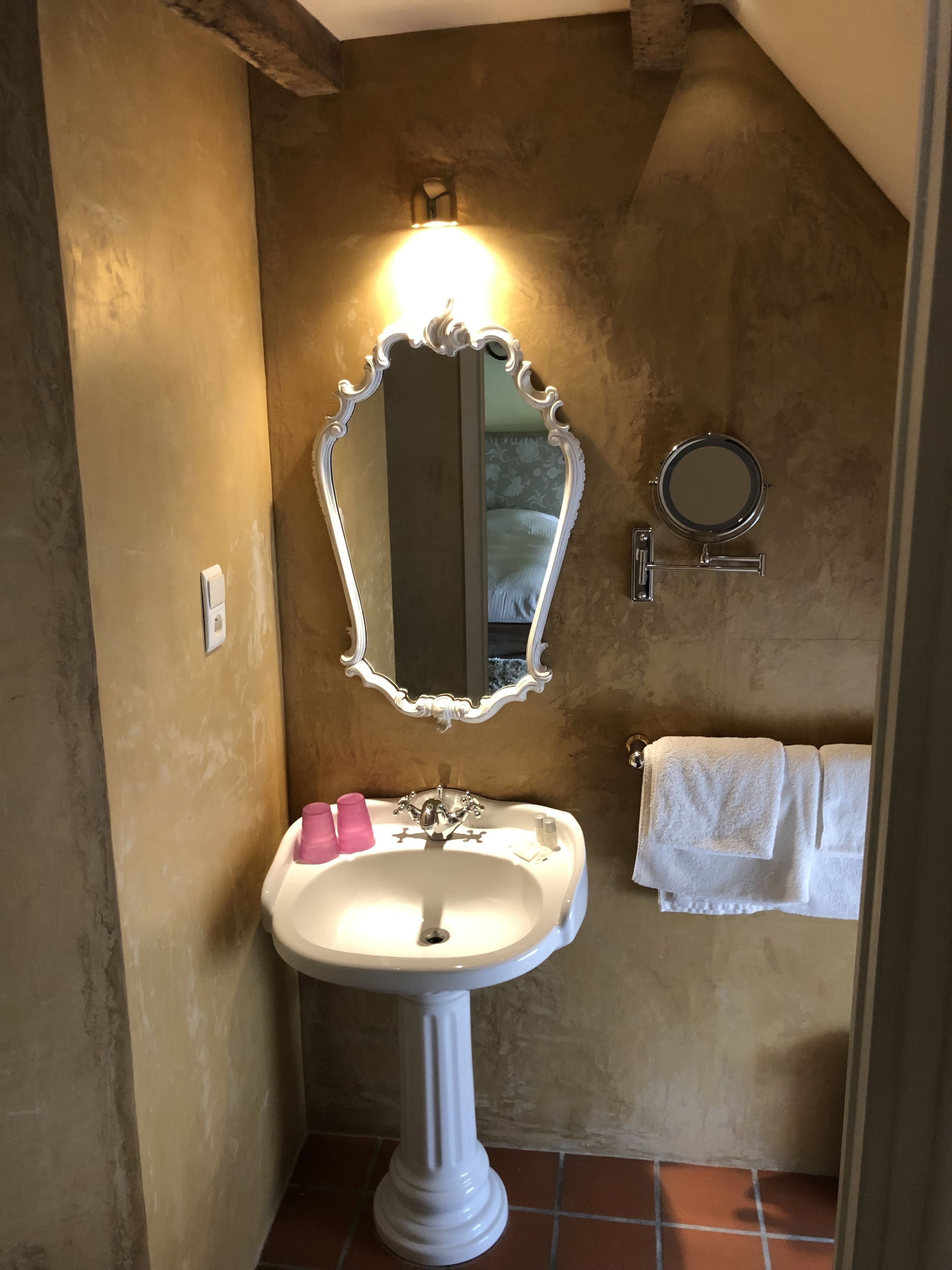 A bathroom with a pedestal sink, a large ornate mirror, a round mirror, and a towel rack.