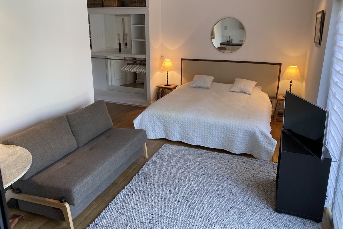 Un dormitorio con cama, un sofá gris, una mesita, un espejo y un armario.