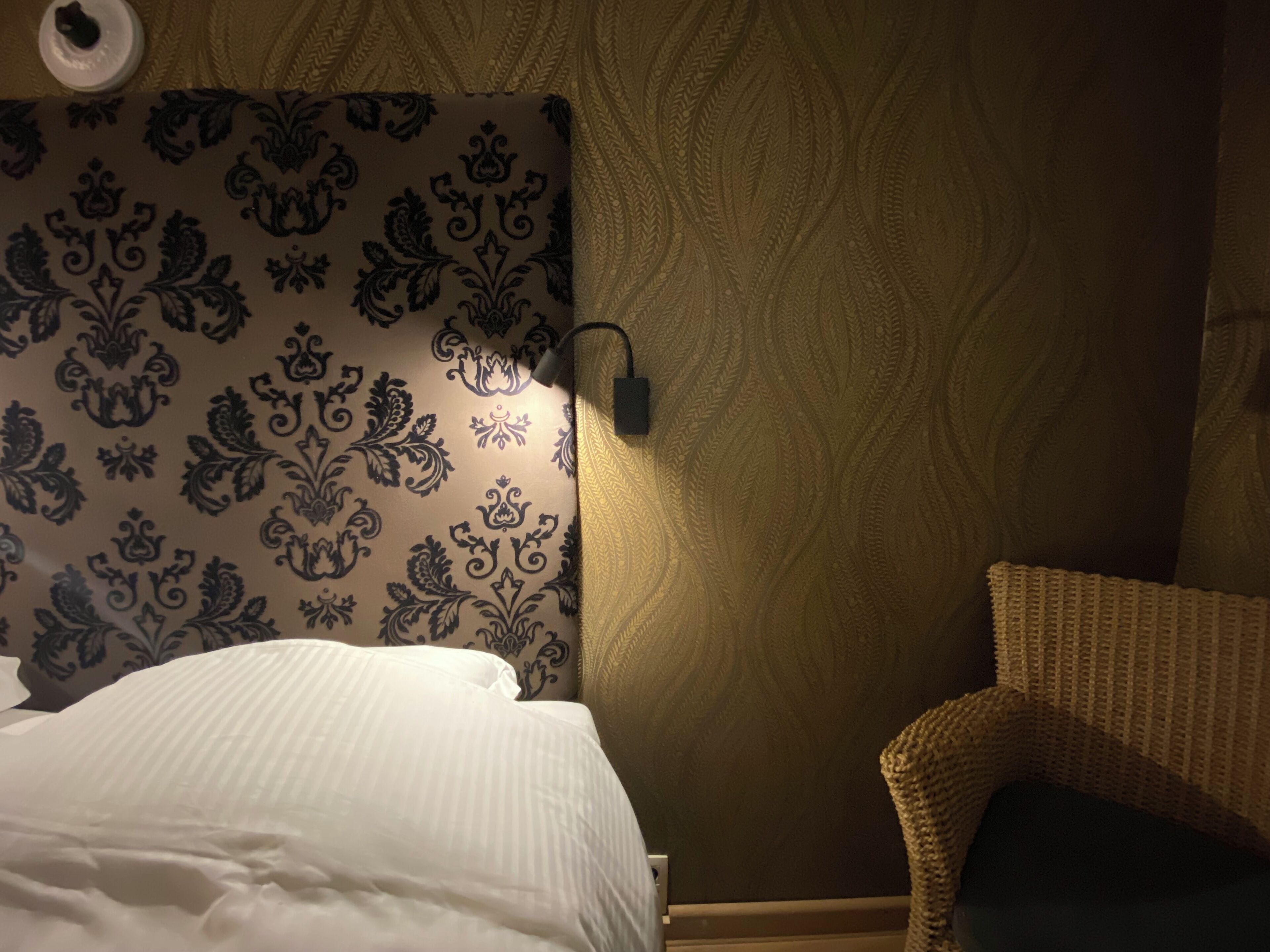 A hotel room with a patterned headboard, a white bed, a wicker chair, and a wall with a textured wallpaper.