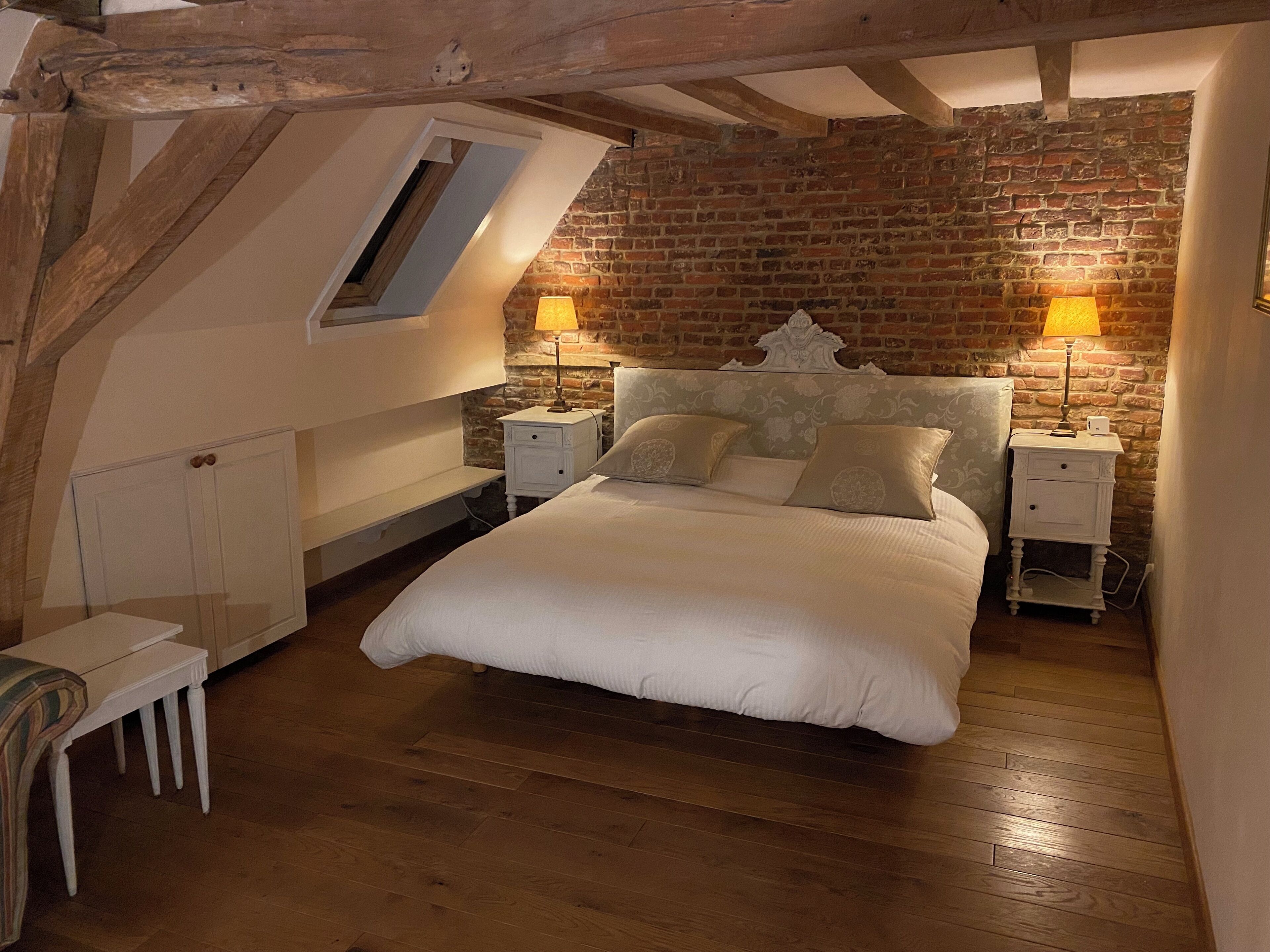 A bedroom with a loft-style ceiling, exposed wooden beams, a brick wall, a bed with white bedding, two bedside tables with lamps, and a small white chair.