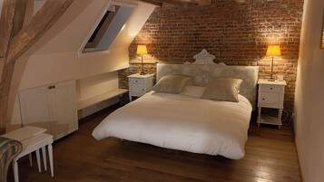 A bedroom with a loft-style ceiling, exposed wooden beams, a brick wall, a bed with white bedding, two bedside tables with lamps, and a small white chair.