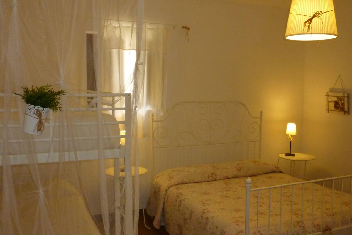 A bedroom with a bed, a bedside table, a lamp, a hanging plant, and a window with curtains.