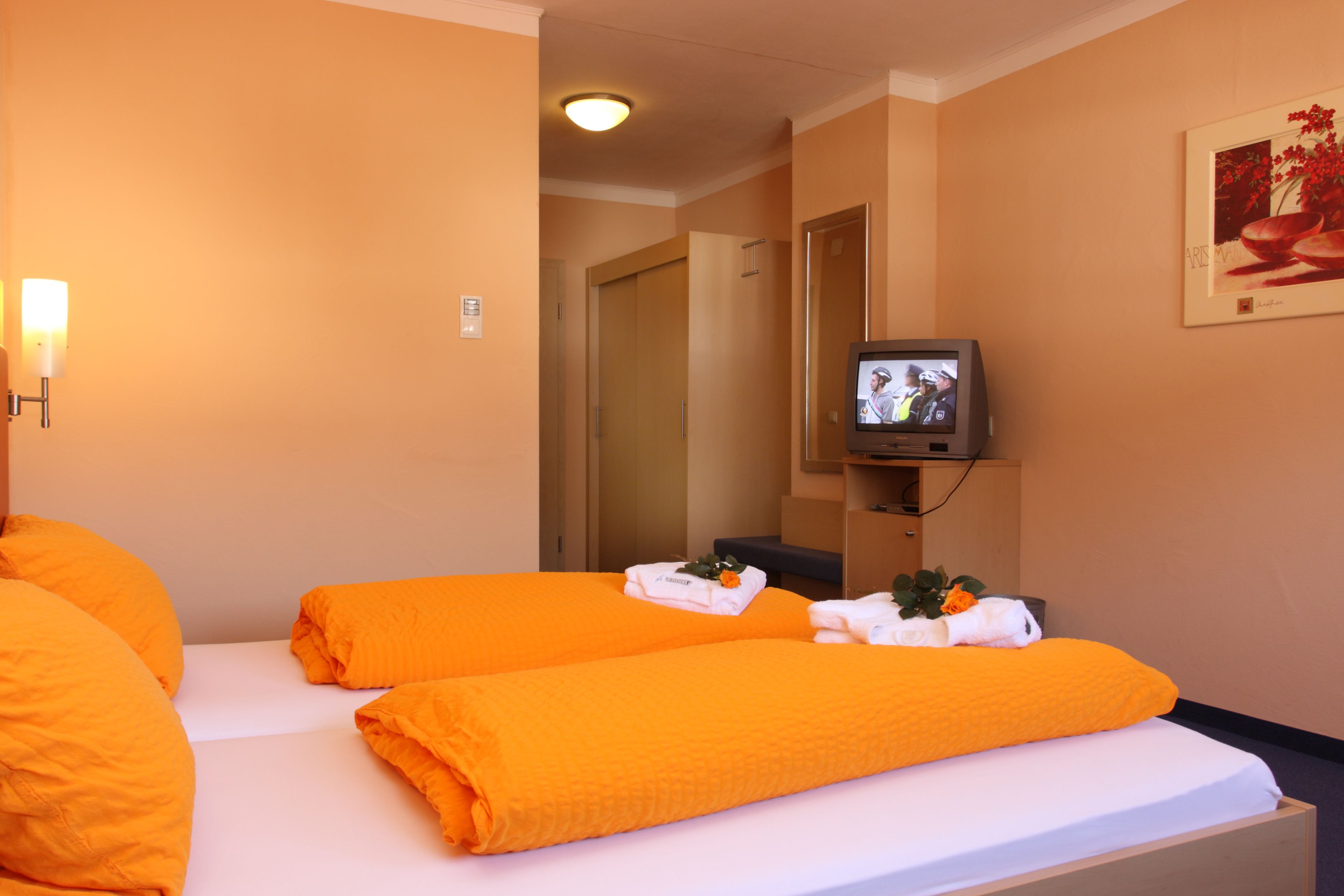 A hotel room with two beds, each with an orange blanket and white pillows. There is a television on a stand to the right of the beds.