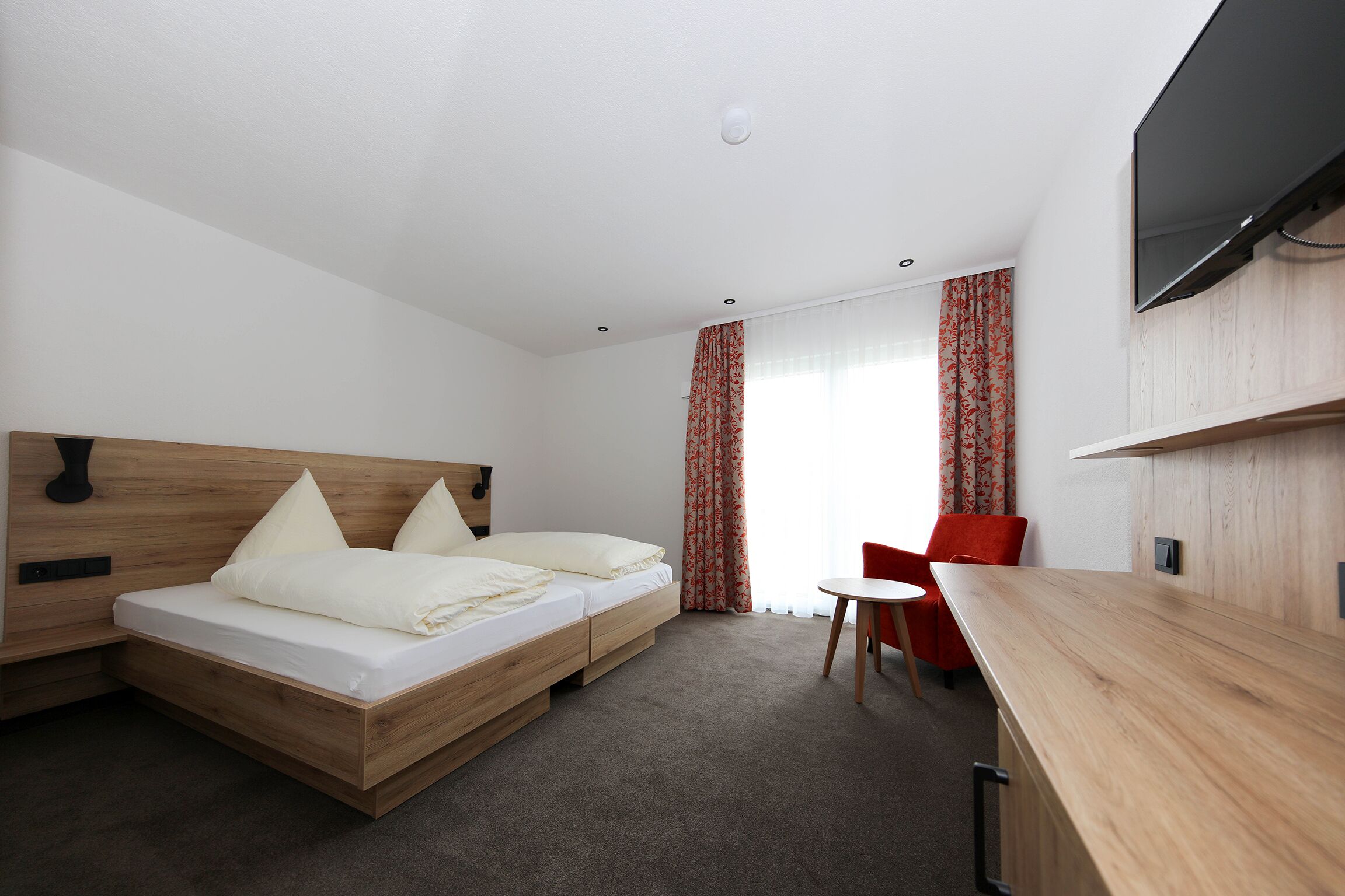 Une chambre d’hôtel moderne avec un lit en bois, un petit bureau, un téléviseur fixé au mur et une fenêtre avec des rideaux.