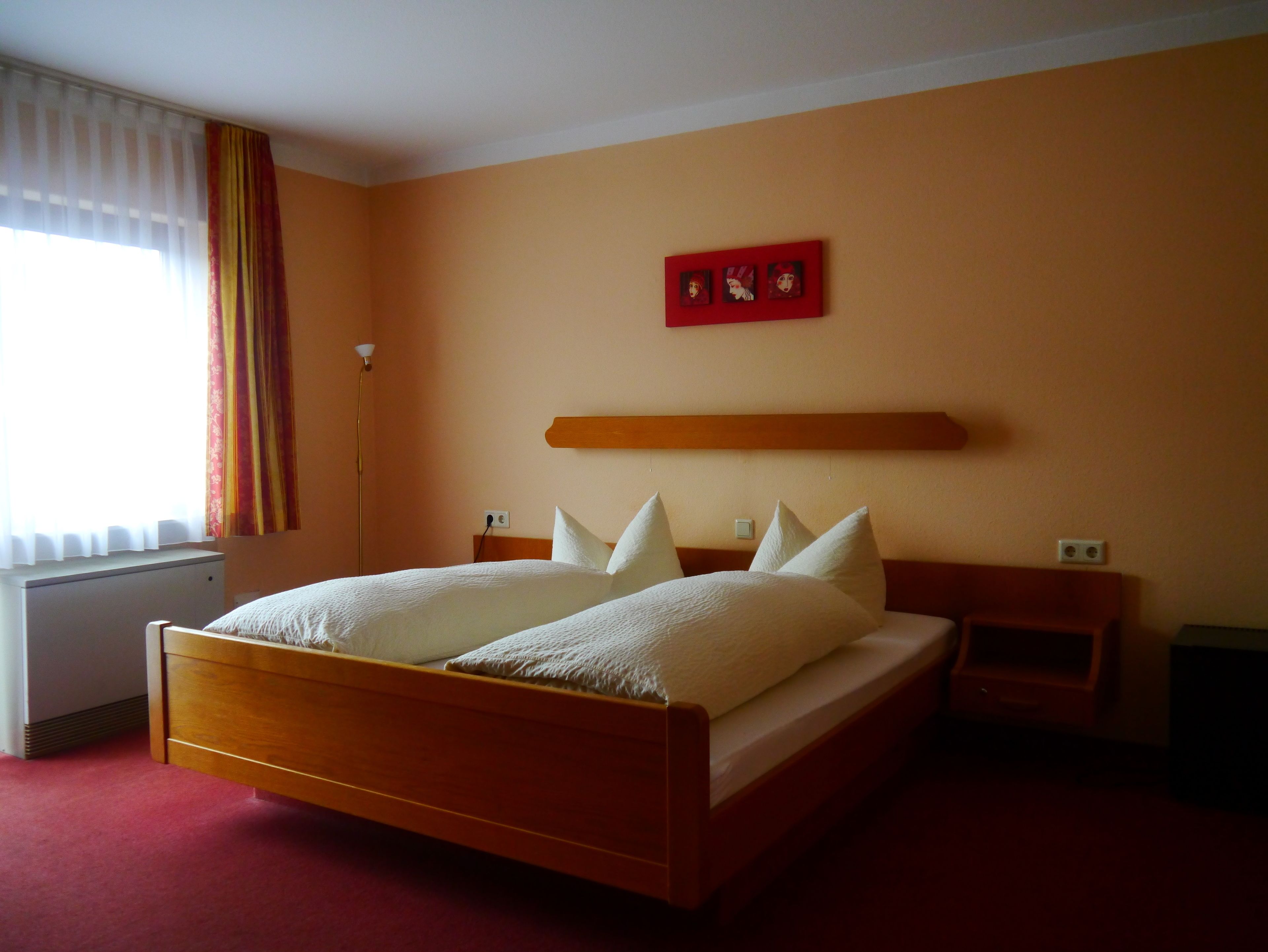 A hotel room with a wooden bed, white bedding, a red artwork above the bed, and a window with curtains.
