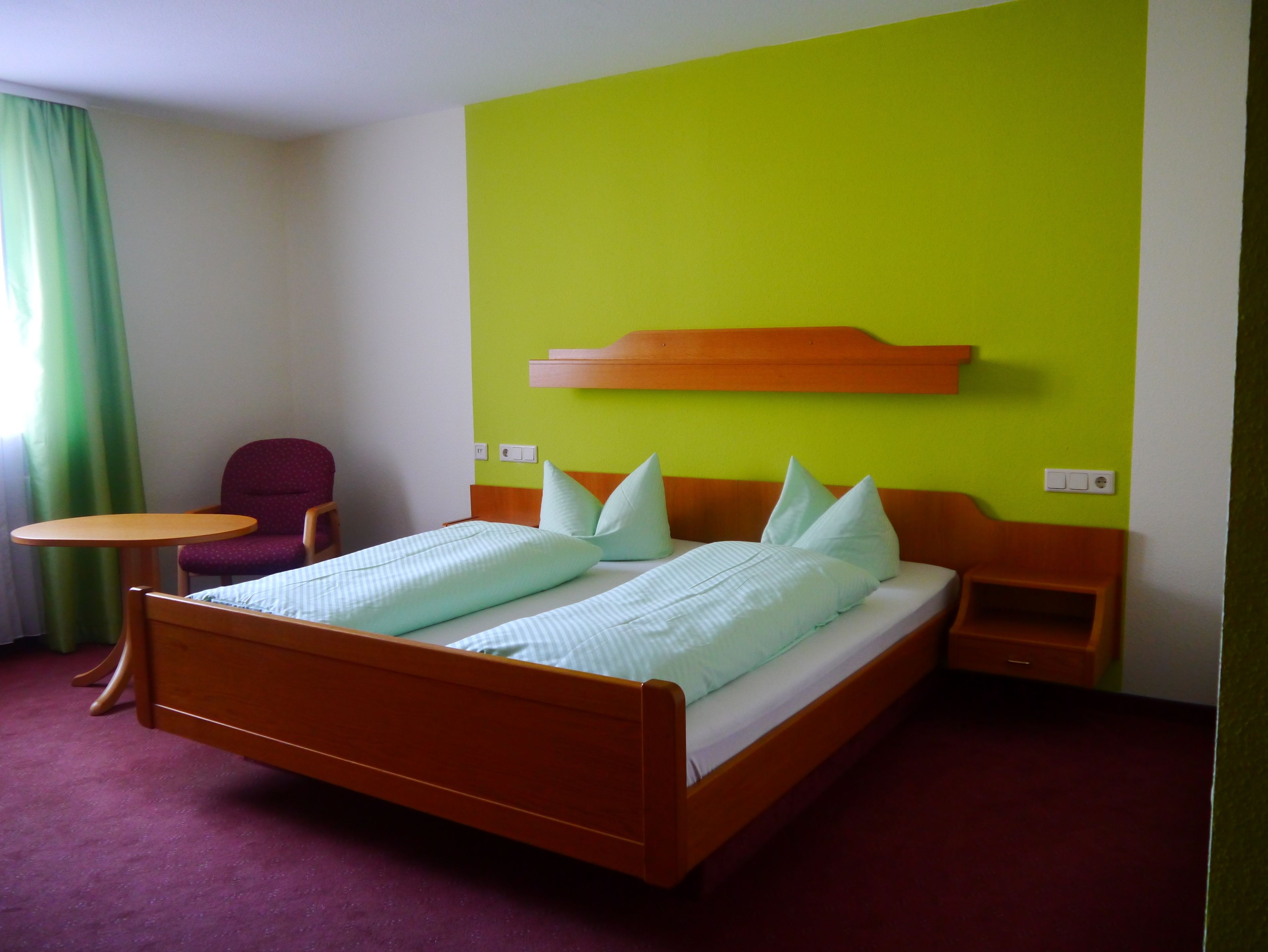 A hotel room with a wooden bed, green walls, a small table, and a chair.