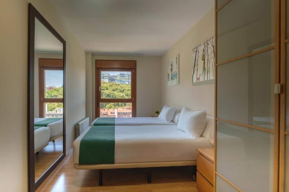 A hotel room with two beds, a large mirror, and a view of greenery and buildings.