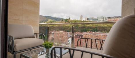 A balcony with two chairs and a small table, overlooking a cityscape with buildings and greenery.
