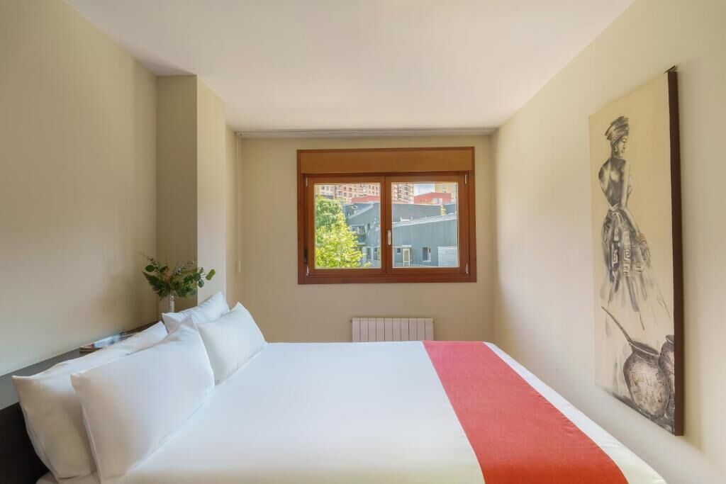 Habitación de hotel con una cama grande, ventana con vista a edificios, radiador y un cuadro en la pared.