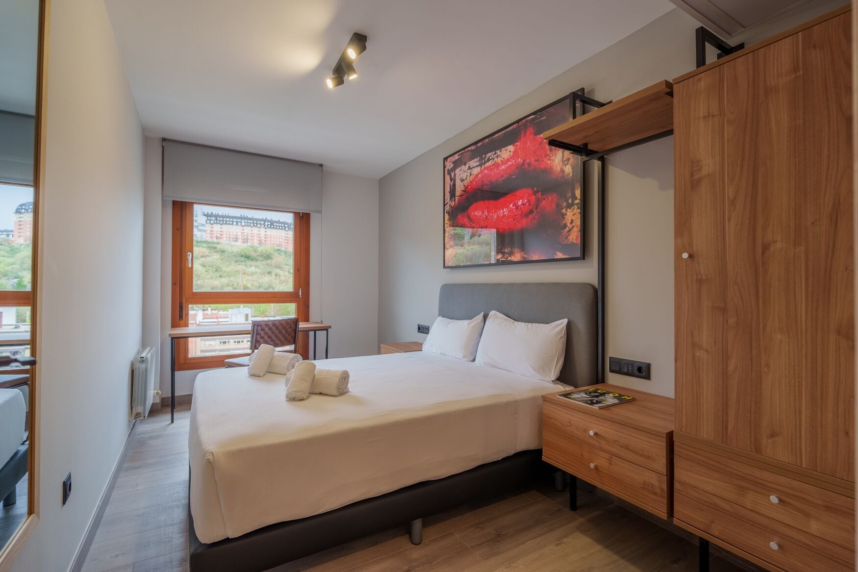 A modern hotel room with a large bed, a wooden wardrobe, a window with a view, and a painting on the wall.