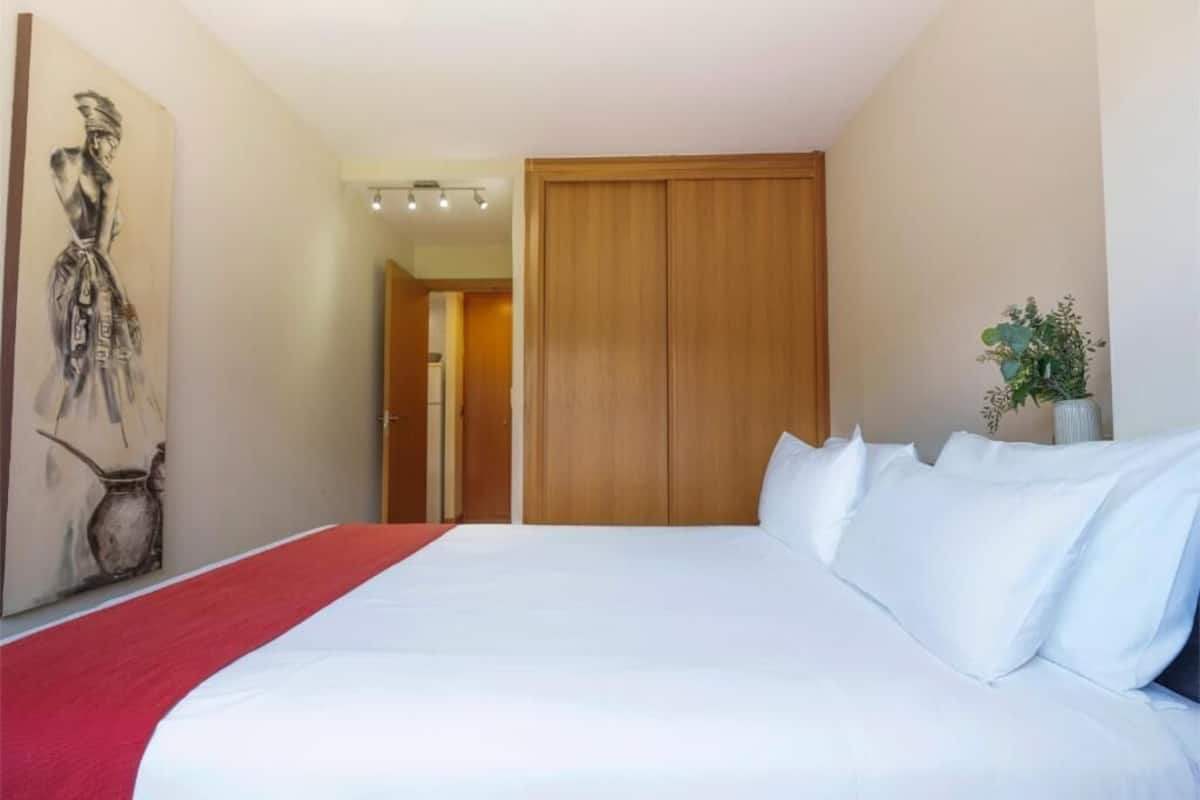 A hotel room with a large bed, a wooden wardrobe, a red throw blanket, and a painting on the wall.