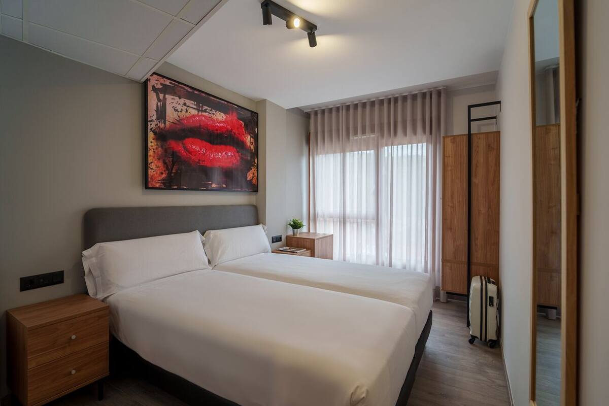 A modern hotel room with a large bed, a nightstand, a small table with a plant, and a large window with curtains.