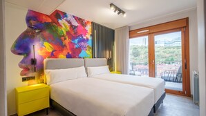 A modern bedroom with two beds, a colorful abstract painting, a yellow nightstand, and a balcony with a view. - Spirit Apartamentos Atxuri (Bilbao)