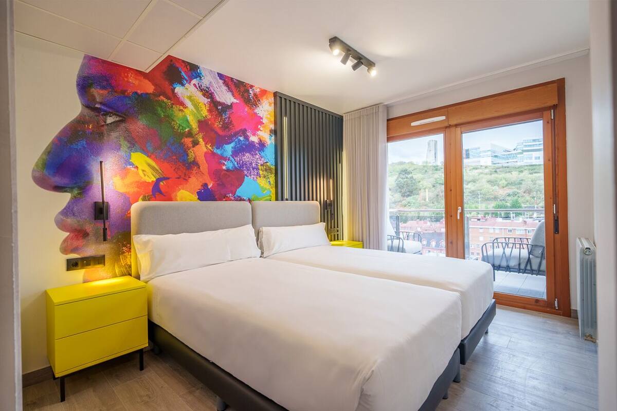 A modern bedroom with two beds, a colorful abstract painting, a yellow nightstand, and a balcony with a view.