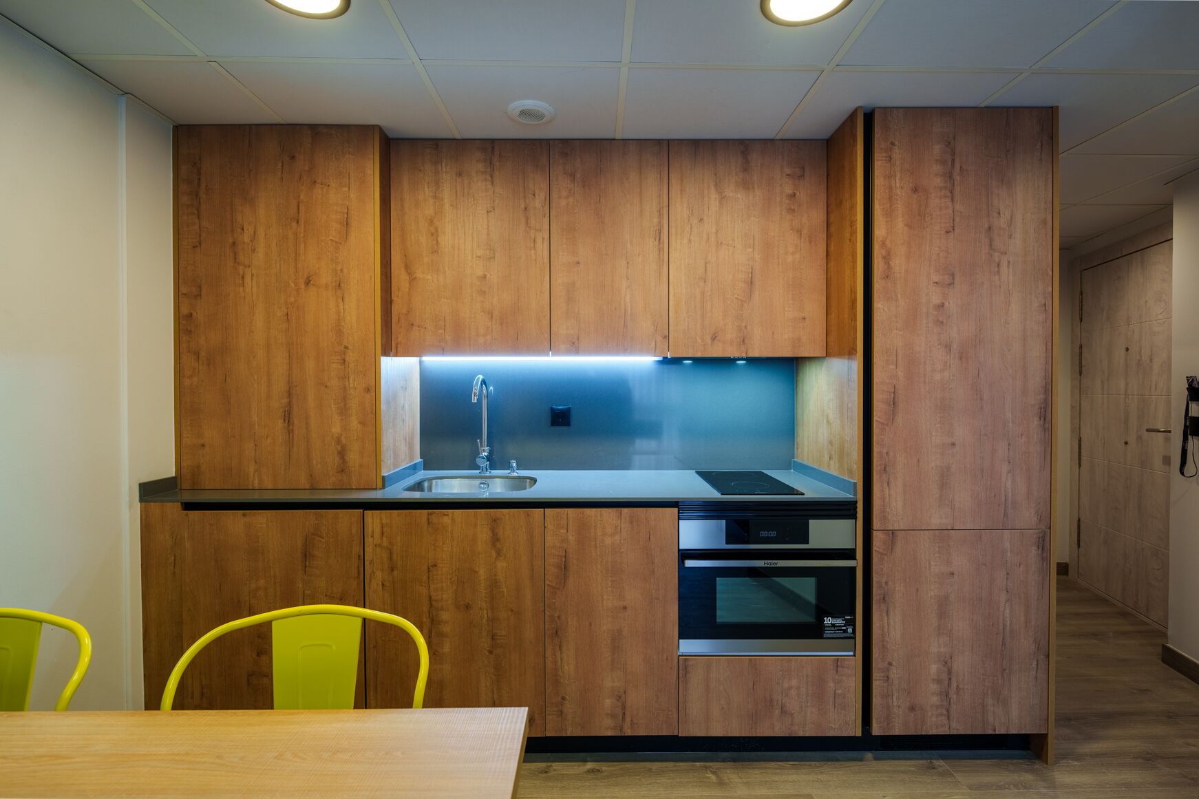 Una cocina compacta con muebles de madera, un lavamanos de acero inoxidable, un horno integrado y una lámpara sobre la encimera muy luminosa.