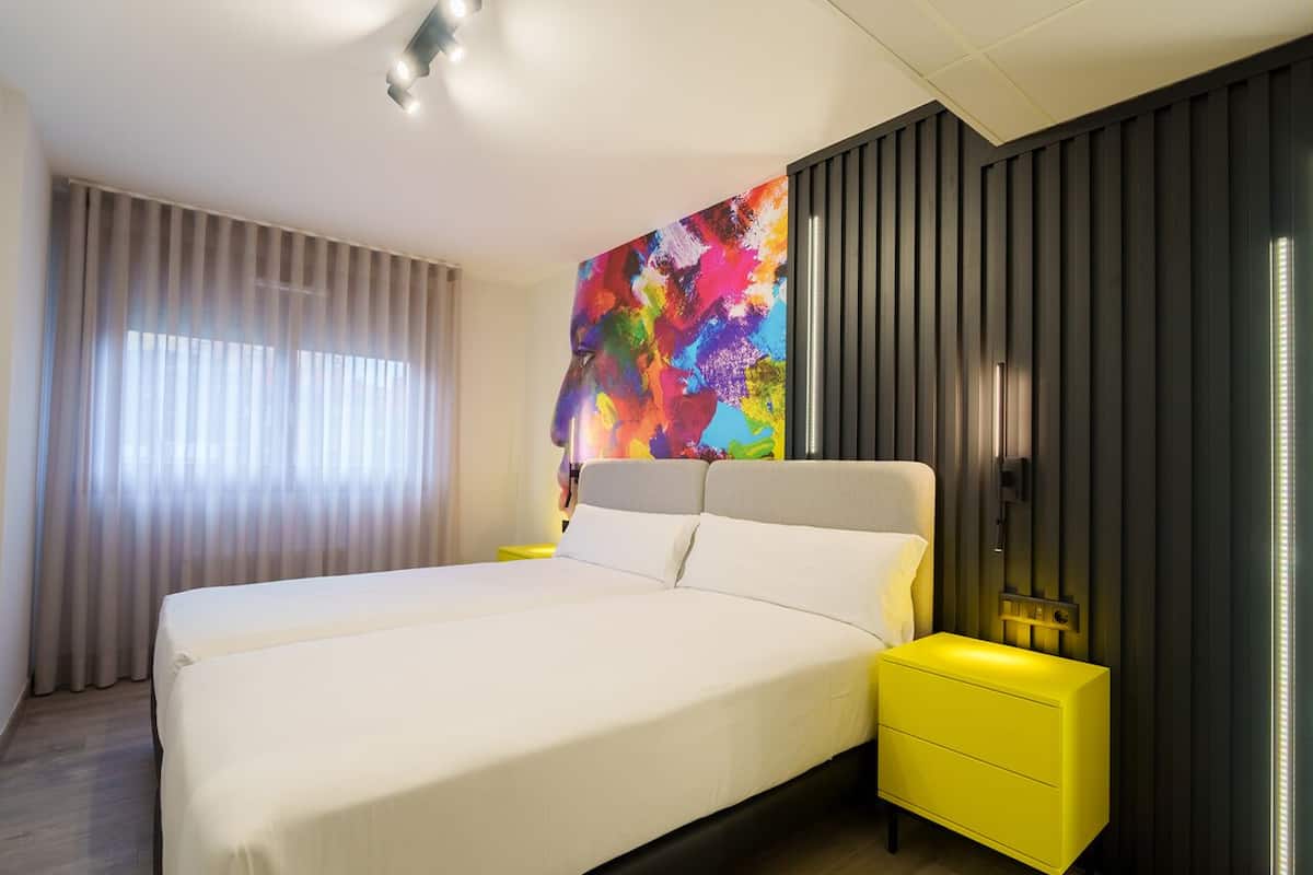 A modern hotel room with a large bed, a bright yellow nightstand, a colorful abstract painting on the wall, and a window with sheer curtains.
