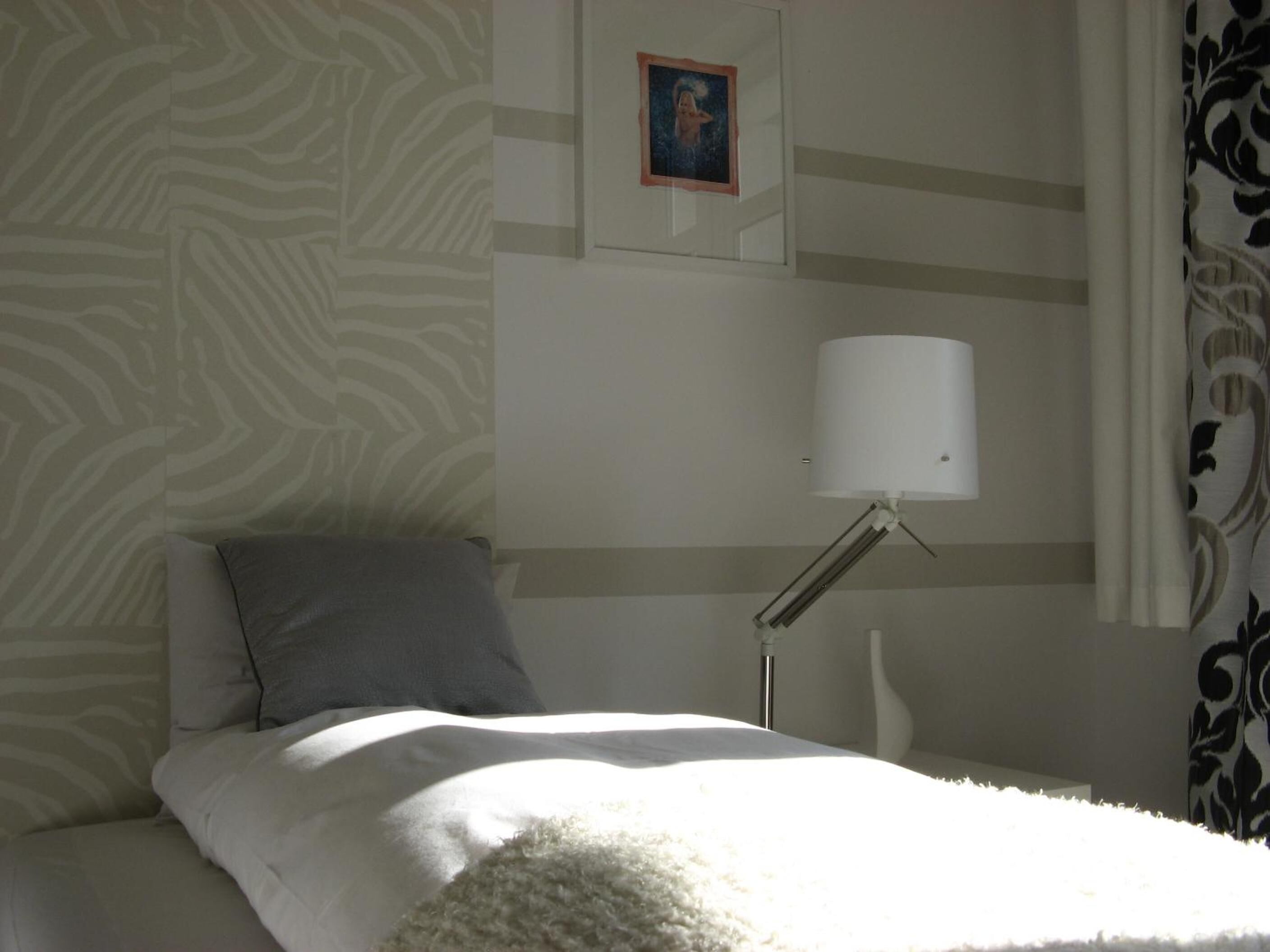 A bedroom with a bed, a pillow, a bedside lamp, a wall with a picture, and patterned curtains.