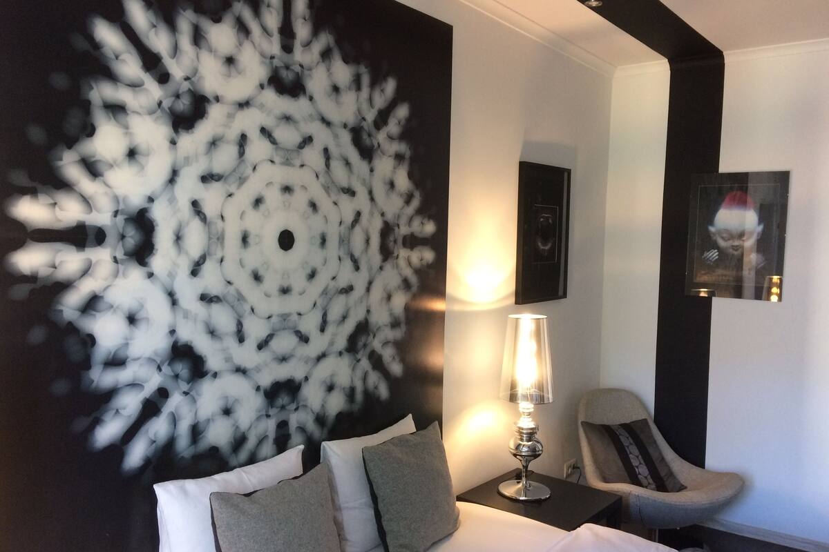 A hotel room with a large abstract wall art, a bed with pillows, a nightstand with a lamp, a chair, and a framed picture on the wall.