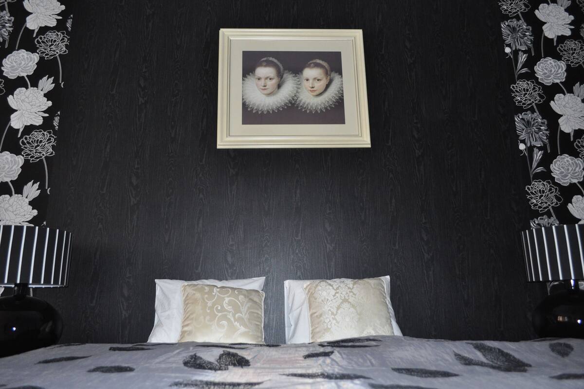 A bedroom with a bed, two pillows, a framed picture on the wall, and floral wallpaper.