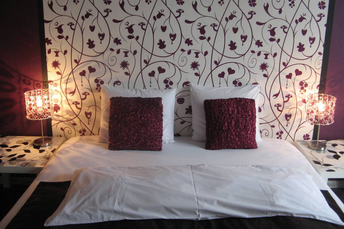 A neatly made bed with white linens and two dark red pillows, set against a wall with a floral pattern.