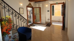 A spacious hallway with a tiled floor, a blue chair, a vase with flowers, and a staircase with a wrought iron railing. - Santa Margherita Guesthouse (Venice)